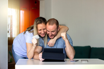 Happy young Caucasian couple shopping online through tablet using credit card at home. Man and women with digital tablet and credit card buying online. Family couple looking at tablet computer at home