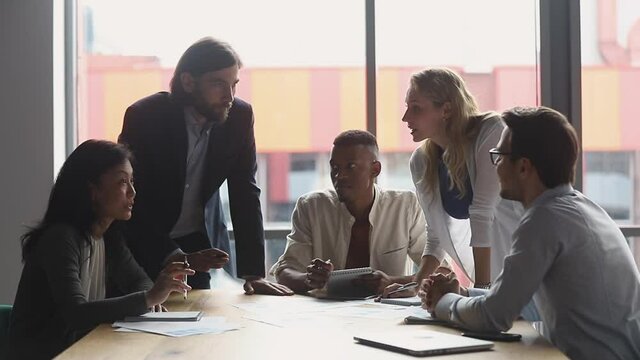 Happy serious friendly multiracial diverse team people brainstorming share ideas talking laughing doing paperwork working in teamwork discuss new design plan at office table at group corporate meeting