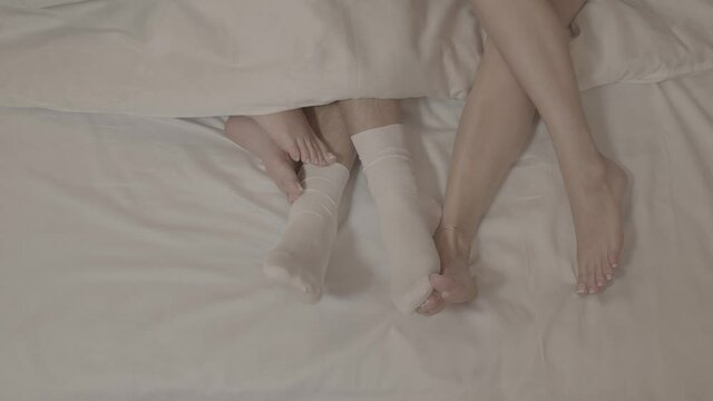 Two women with bare feet laying on bed touching man legs in white socks under blanket on white bed sheets in close up
