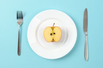 Half of ripe juicy apple on a white dinner plate, table knife and fork on a blue background. Concept of healthy eating with fiber and vitamins, vegetarianism and raw food diet.