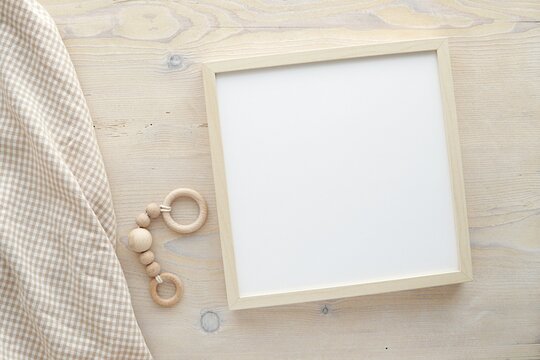 Wooden square frame mockup, empty frame mock up for nursery, baby room artwork, photo, sayings, flat lay composition with baby blanket and baby teether on wooden background.