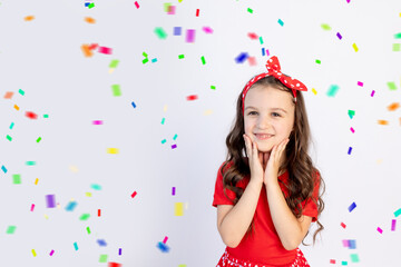 Obraz na płótnie Canvas A cute little girl in a red dress on a white isolated background with a streamer folded her hands on her cheeks. Space for text. Little girl celebrates birthday, holiday concept