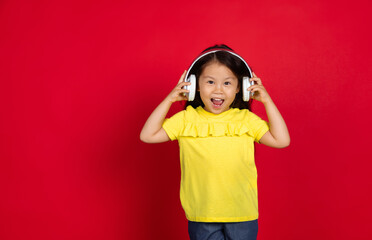 Listening to music, dancing. Beautiful little girl on red background. Half-lenght portrait of happy child. Cute asian girl in yellow wear. Concept of facial expression, human emotions, childhood.