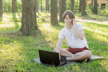 Young man is sitting on the grass in park with his laptop and talking on the phone. Distance learning concept.