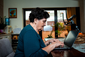 Fototapeta na wymiar An elderly senior lady sits at a dining table and uses a computer. She looks at something on the Internet and intends to order some things through the online store
