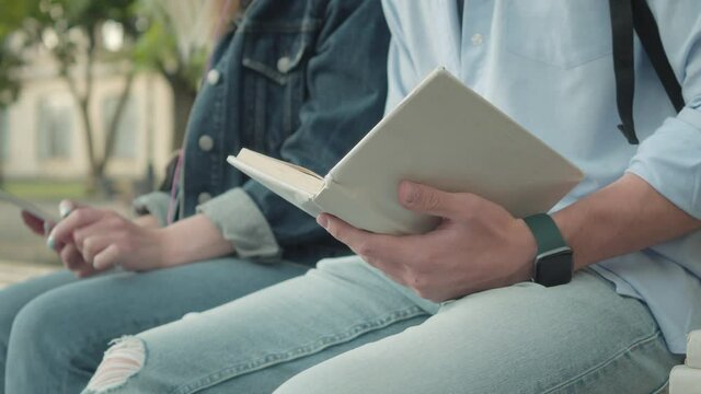 Close-up of unrecognizable young man sitting with book outdoors and young girl holding smartphone at the background. Two university students studying in college yard. Education concept.