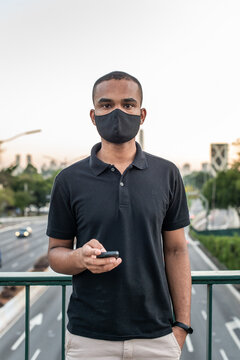Jovem negro parado na rua olhando para frente com celular na m&atilde;o usando m&aacute;scara de prote&ccedil;&atilde;o, S&atilde;o Paulo