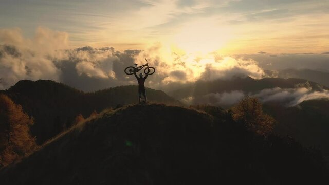 Aerial - Happy man holding his bicycle above his head at sunset after a mountain biking trip to the beautiful mountains. 4K