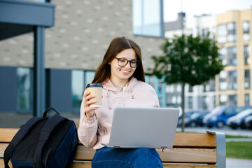Fototapeta premium Positive girl in jeans with coffee cup in hand sits on a bench and working at a laptop. Freelance work concept