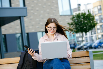 Positive girl in jeans with a mobile phone in hand sits on a bench and working at a laptop. Freelance work concept