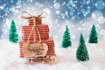 Sled With Christmas Gift And Label With German Frohe Weihnachten Und Ein Glueckliches 2021 Means Merry Christmas And A Happy 2021. Snow With Christmas Tree And Blue Sparkling Background