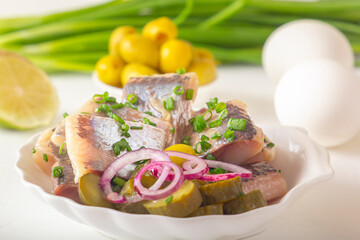 Slices of pickled herring with red onion, fermented cucumber, fried potatoes, olives, boiled eggs and herbs on a light wooden table. Close-up