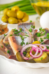 Slices of pickled herring with red onion, fermented cucumber, fried potatoes, olives, boiled eggs and herbs on a light wooden table. Close-up