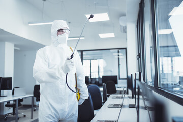 Office disinfection during COVID-19 pandemic. Man in protective suit and face mask spraying for disinfection in the office