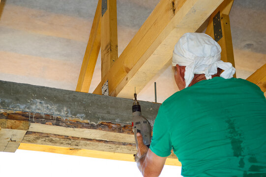 A Man Stands With His Back In A White Kerchief On His Head And A Green T-shirt Works At A House Construction Site, Drills A Wooden Beam With A Drill; Sweaty From Active Work