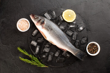 Fresh sea bass with ice cubes,rosemary,lemon,sea salt and dried pepper on black background.Top view.