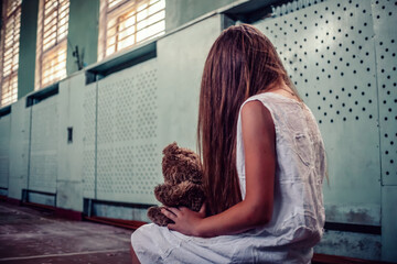 girl in a white dress with long hair sits in an abandoned gym of a destroyed school and holds a Teddy bear in her lap, a photo from the back. concept of horror, mysticism