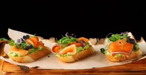 bruschetta with salmon closeup. tapas with salmon on guacamole and topped microgreen on wooden board and black background