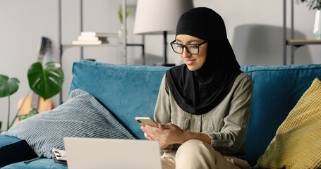 Portrait of beautiful happy arab girl in hijab working at home using smartphone and laptop computer while sitting on sofa.