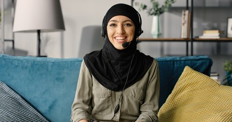 Portrait of cheerful muslim young woman at home, working remotely while sitting on couch in living...