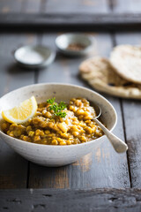 Vegan friendly lentil daal served in hand thrown pot, garnished with lemon wedges and herbs on grey wooden table