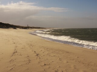 Landscape on the island of Sylt northern tip, in the foreground fine sandy beach and dunes in stormy winter weather , in the background a red - white streaked lighthouse, wavy sea whipped by the wind