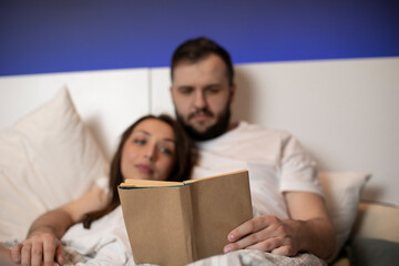 couple of lovers wearing white pajamas reading one book for two in bed together. Togetherness concept. Happiness, serenity, harmony concept