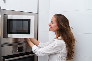Fototapeta na wymiar Portrait of a beautiful young modern long haired smiling woman cooking in the microwave oven in domestic kithen indoor.