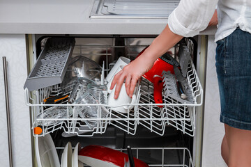 Dishwasher with an open door and extended basket shelves with clean dishes and a lovely woman in a short skirt taking out or loading a mug from the dishwasher on the white facade of a standard kitchen