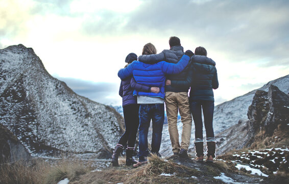 Rear view of four people standing arm in arm on a mountain, snow-capped peaks in the distance.