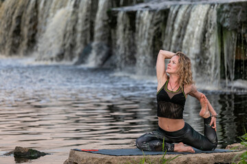 Blonde woman in sportswear practicing yoga on a riverside in front of a waterfall. Girl laid fitness mat on a rock and doing a stretching exercise outdoor in harmony with nature.