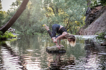 Fototapeta na wymiar Young woman practicing yoga outdoors in harmony with nature. Fitness girl balancing on arms a small rock in the middle of a forest river