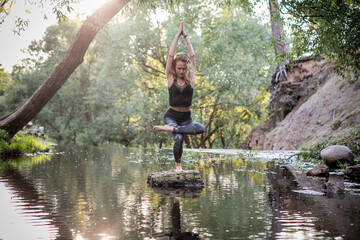 Naklejka na ściany i meble Young woman practicing yoga outdoors in harmony with nature. Fitness girl balancing on one leg a small rock in the middle of a forest river with hands clasped over her head