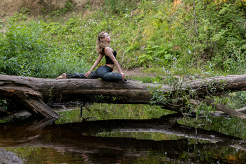 Blonde woman in sportswear practicing breathing yoga on a log over the forest river. Fitness girl doing a stretching exercise outdoor in harmony with nature.