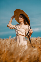 Obraz premium Attractive and beautiful girl in a beige midi dress with a big straw hat, in a wheat field