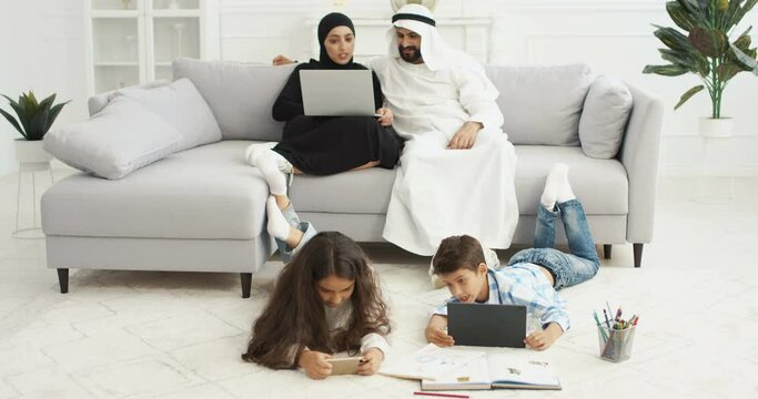 Arabian happy kids lying on floor and playing on gadgets. Small sister and brother using smartphone and tablet. Couple of parents sitting on couch with laptop computer. Arabs mother and father rest.