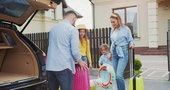Summer family vacation. Cheerful handsome parents packing and closing up car trunk preparing for roadtrip. Excited kids staying in sunny street. Concept of lifestyle, happy family, travel together.