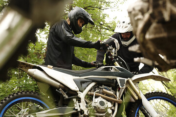 Male motorcyclists in helmets preparing motorcycle for forest race or checking it after fall