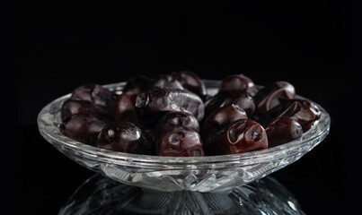 Crystal saucer with a bunch of dried dates on a dark mirror background.
