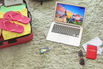 Open page of online booking service on screen of laptop and suitcase with belongings at home