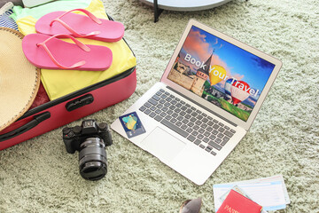 Open page of online booking service on screen of laptop and suitcase with belongings at home