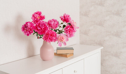 A large bouquet of peonies in a pink vase in a bright room on a white chest, books are nearby. fresh flowers at home, greenhouse, minimalist, cozy loft interior