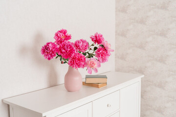 A large bouquet of peonies in a pink vase in a bright room on a white chest, books are nearby. fresh flowers at home, greenhouse, minimalist, cozy loft interior