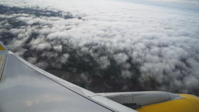 POV Airplane Passenger Window: White Blanket And Billowing Clouds Below With View Of Aircraft Wing And Yellow Engine In Flight, Handheld Slow Motion