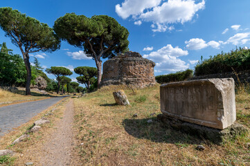Fototapeta na wymiar View of the circular brick tomb and epigraph on the Via Appia on a sunny day. The road was the most important of the Roman Empire, remains of other tombs, summer day, maritime pines, cypresses.