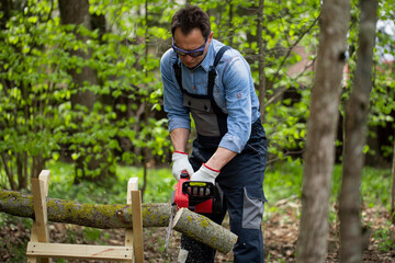 Brutal adult forest worker in overalls saws tree trunk with the chainsaw on sawhorses in forest....