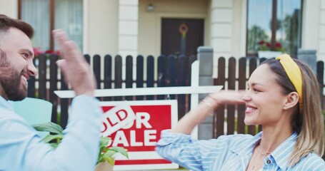 Joyful happy young couple standing in front of a sold sign looking at their newly purchased home. Family moving to a new apartment area at sunny day. Husband gives five to the wife.