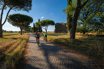 A stretch of the Appian Way, one of the most important streets of the Roman Empire. Dense vegetation, maritime pines, two cycles next to a tumulus grave on a summer day. Rome, Italy.