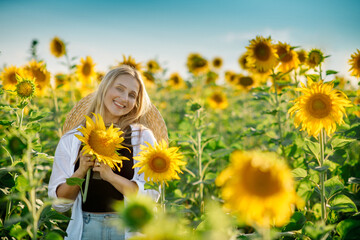 Fototapeta na wymiar Beautiful young blonde woman in a hat and white shirt walks and laughs in a sunflower field 
