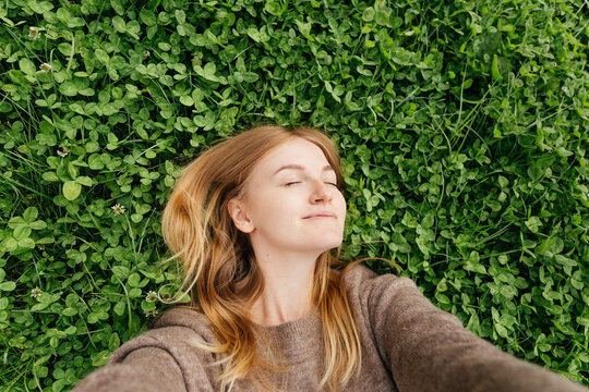 Beautiful young hipster woman taking selfie on green grass at the park. Clover in meadow in spring. Travel and active life concept. Smiling dreaming woman lying on the grass